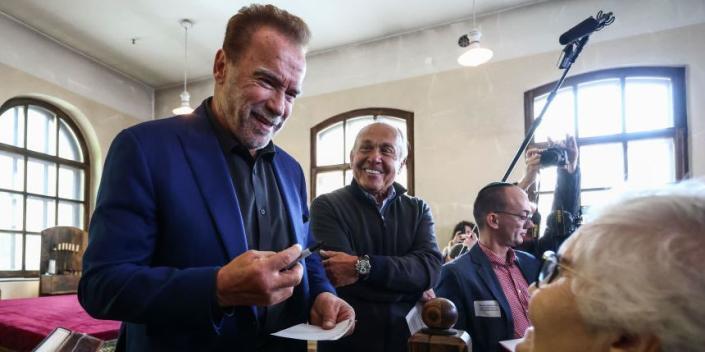 arnold schwarzenegger visits auschwitz memorial and smiles at an older woman as other people look on and take photos