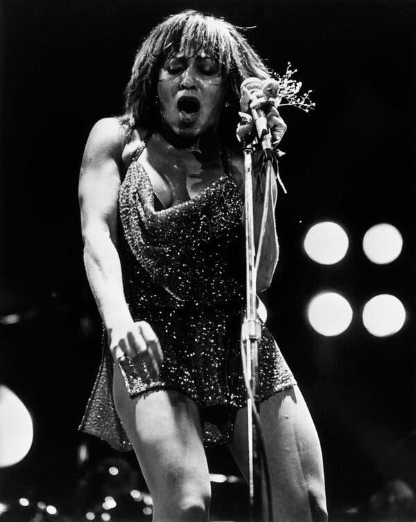In a black-and-white photograph, Tina Turner is wearing a sparkly dress and gripping a microphone on a stand, her mouth open.