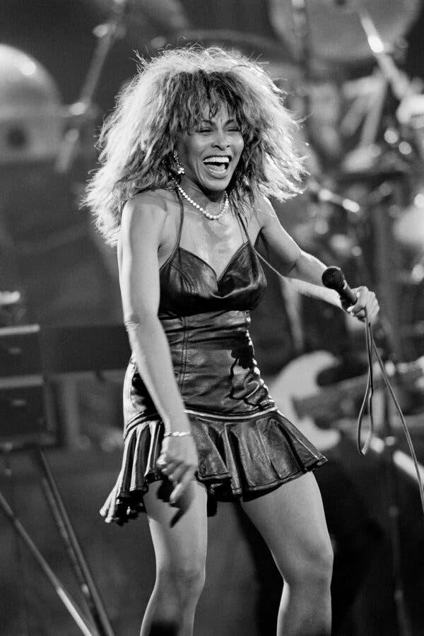 Tina Turner in a black and white photo, standing onstage in a leather dress, holding a microphone, with a big smile on her face.