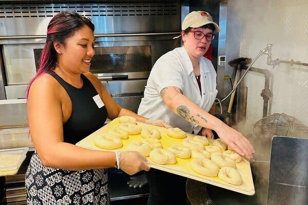 A person with long black hair with pink highlights in a black tank top and skirt is holding a platter of uncooked bagels. Another person, in chef’s whites, red glasses and a beige baseball hat, is helping to push them into a cooker.